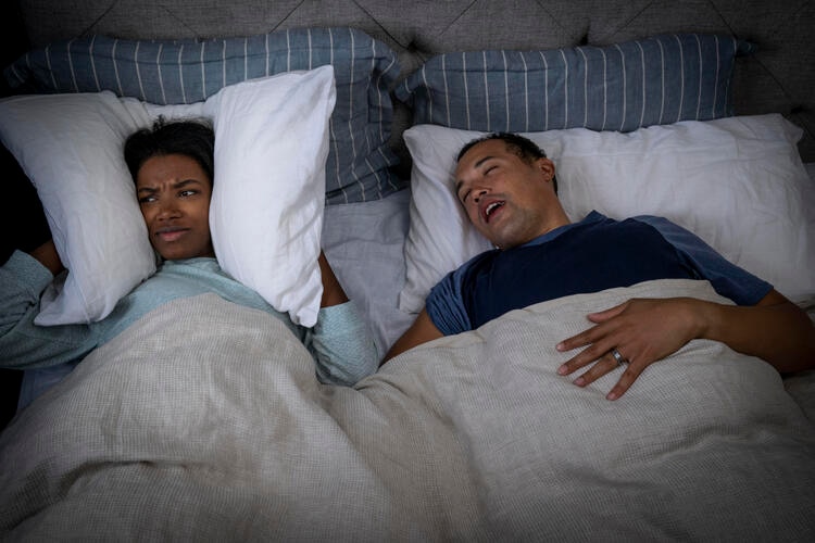 Ein Mann liegt schnarchend im Bett, während seine Partnerin sich die Ohren mit einem Kissen zuhält.